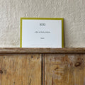 Luxury white IOU gift card with dark green text on a textured surface, in front of a lime green envelope placed on a wooden shelf, with a cream wall behind it.