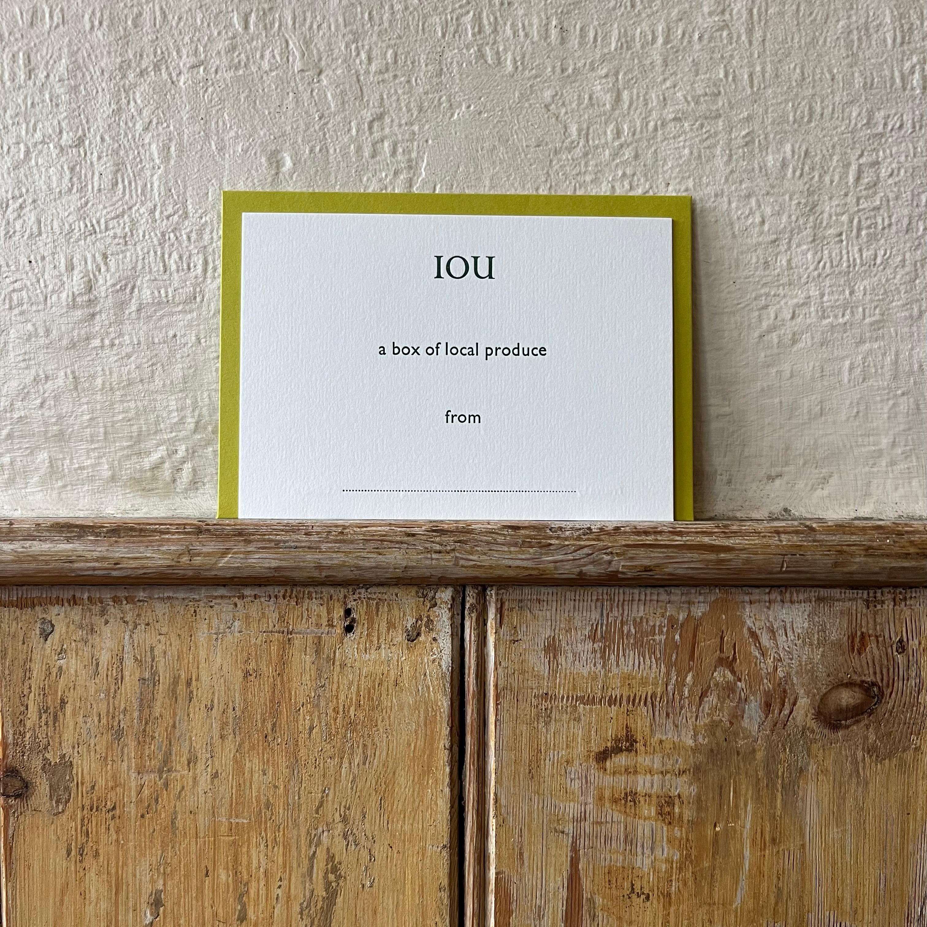 Luxury white IOU gift card with dark green text on a textured surface, in front of a lime green envelope placed on a wooden shelf, with a cream wall behind it.
