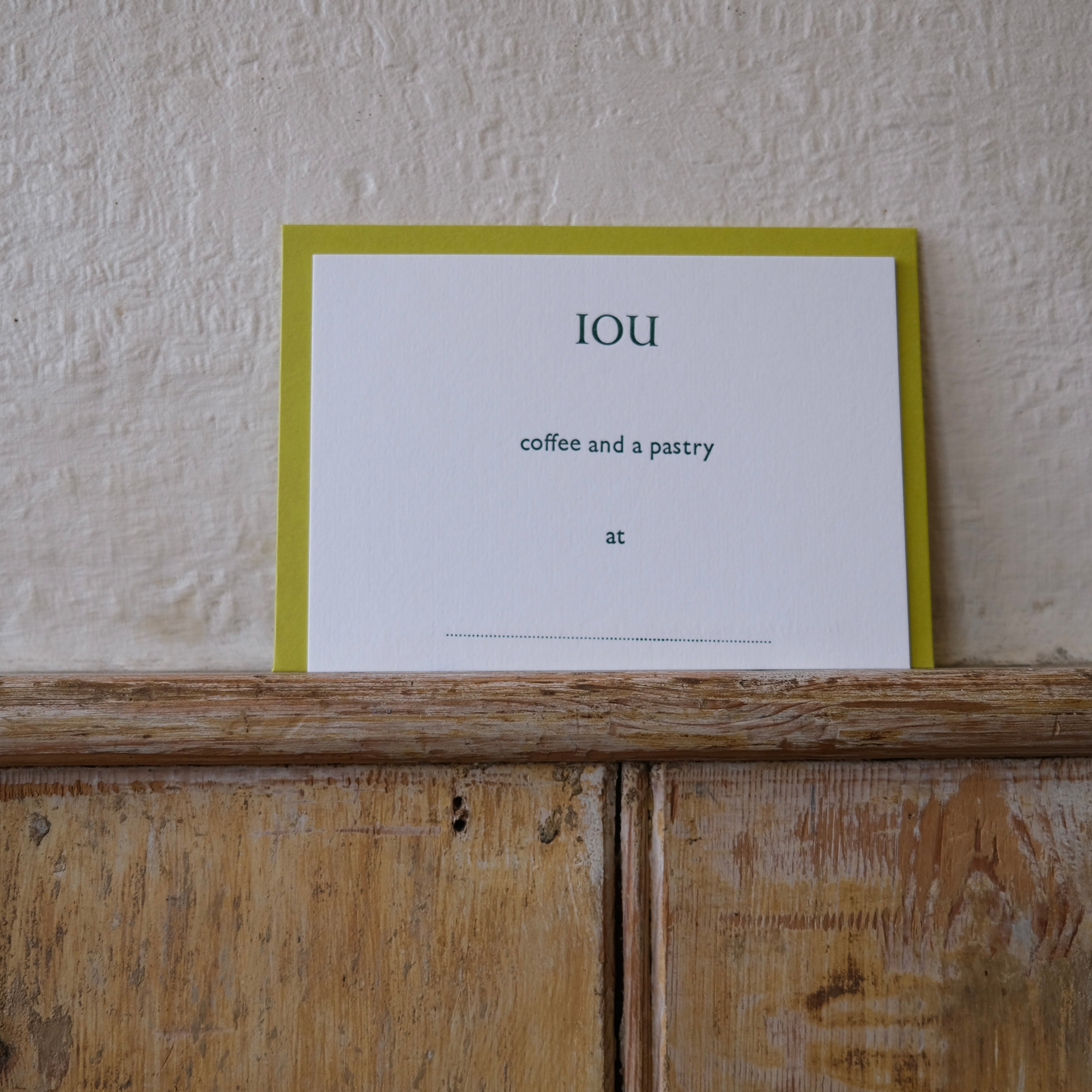 Luxury white IOU gift card with dark green text on a textured surface, in front of a lime green envelope placed on a wooden shelf, with a cream wall behind it.