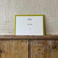 Luxury white IOU gift card with dark green text on a textured surface, in front of a lime green envelope placed on a wooden shelf, with a cream wall behind it.