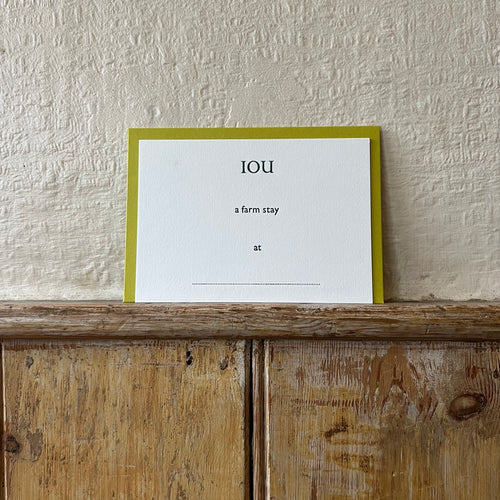 Luxury white IOU gift card with dark green text on a textured surface, in front of a lime green envelope placed on a wooden shelf, with a cream wall behind it.