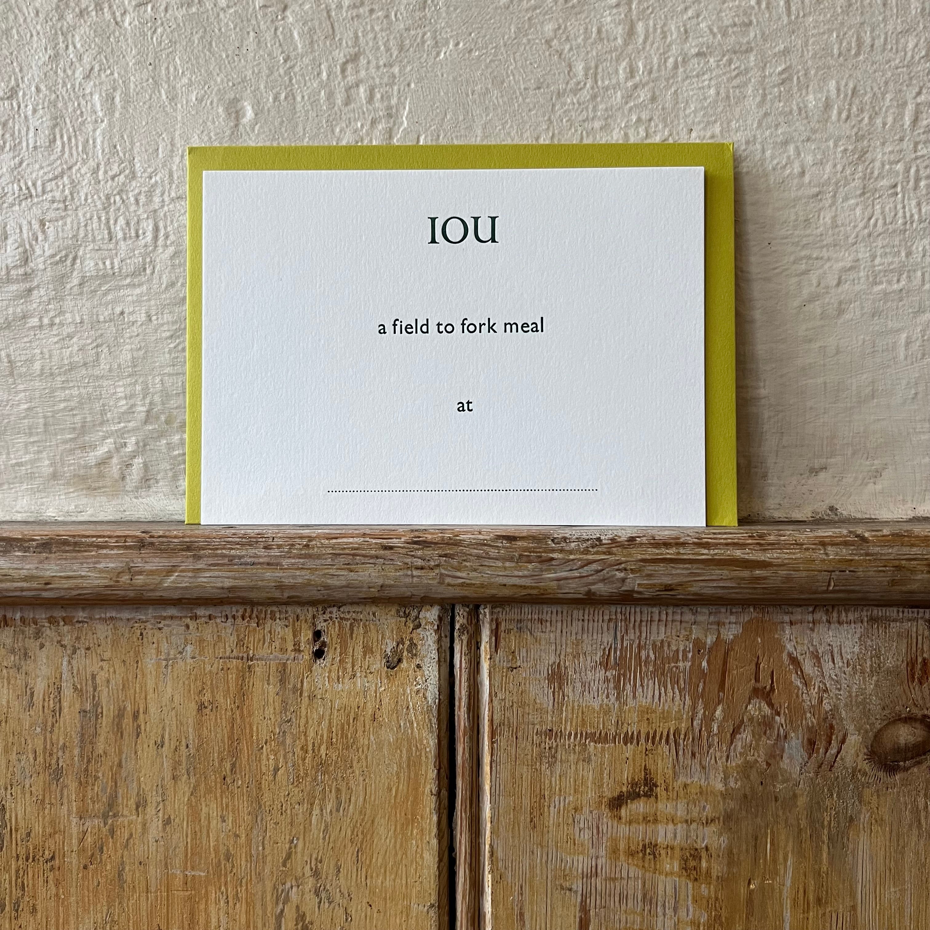 Luxury white IOU gift card with dark green text on a textured surface, in front of a lime green envelope placed on a wooden shelf, with a cream wall behind it.