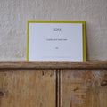 Luxury white IOU gift card with dark green text on a textured surface, in front of a lime green envelope placed on a wooden shelf, with a cream wall behind it.