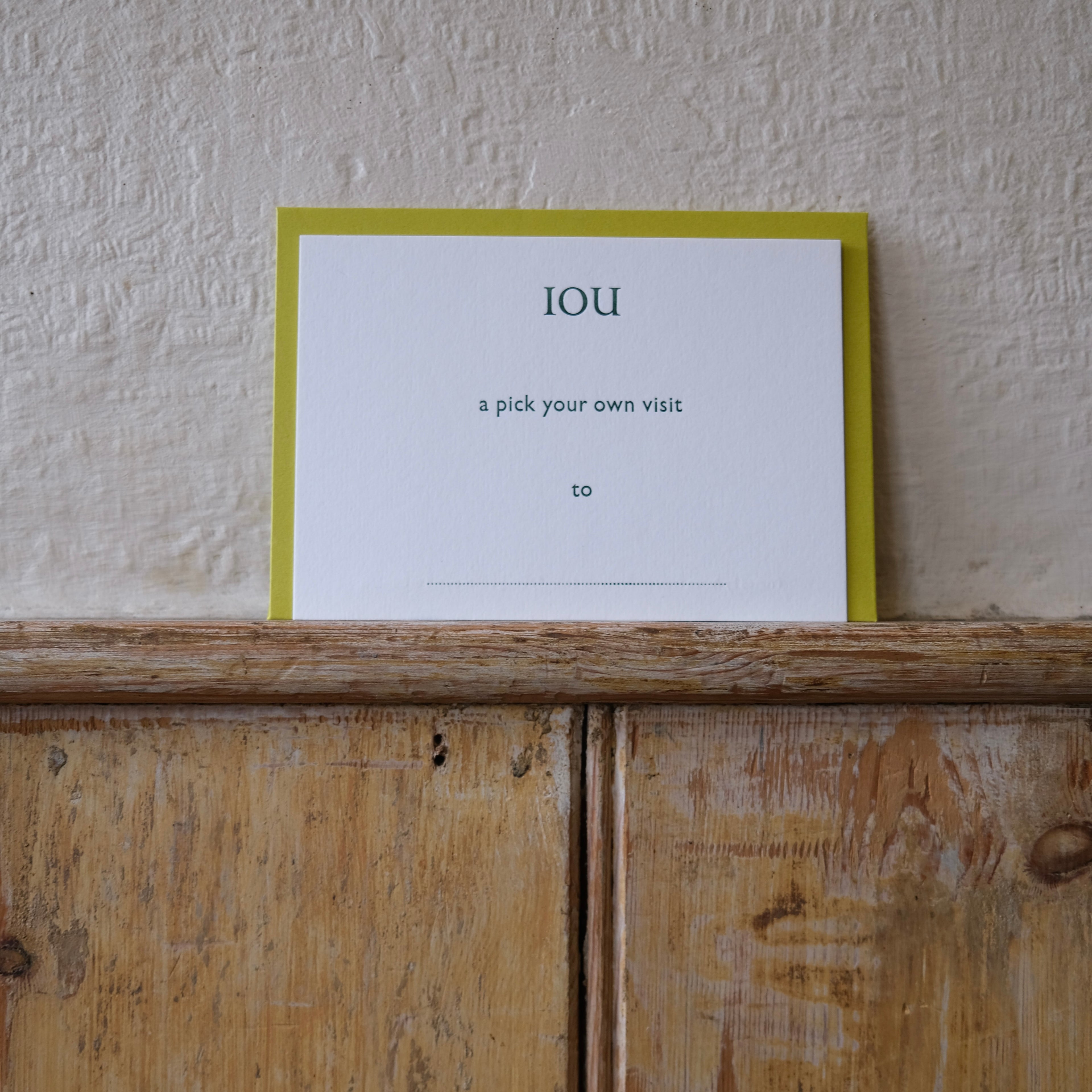 Luxury white IOU gift card with dark green text on a textured surface, in front of a lime green envelope placed on a wooden shelf, with a cream wall behind it.