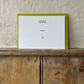 Luxury white IOU gift card with dark green text on a textured surface, in front of a lime green envelope placed on a wooden shelf, with a cream wall behind it.