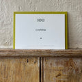 Luxury white IOU gift card with dark green text on a textured surface, in front of a lime green envelope placed on a wooden shelf, with a cream wall behind it.
